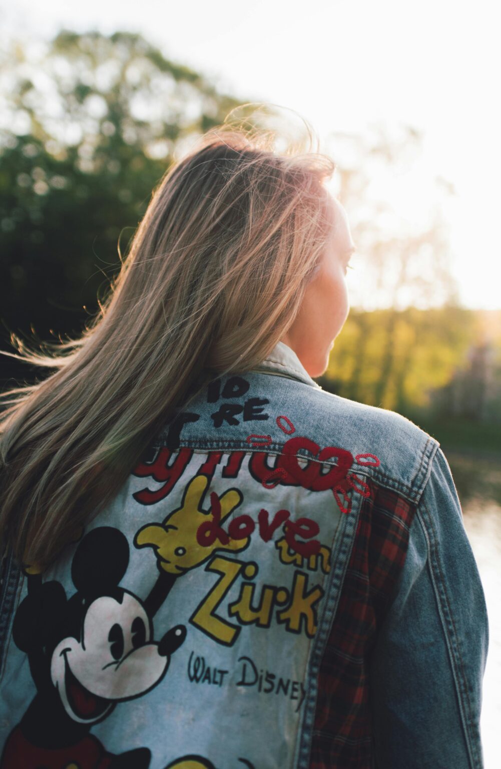 A woman with long blonde hair wearing a denim jacket featuring a colorful Mickey Mouse design in an outdoor setting.