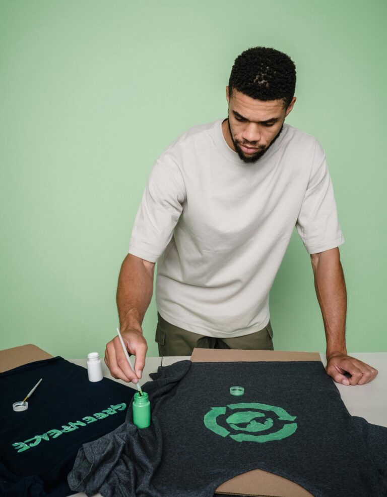 Man with beard painting custom designs on t-shirts in a studio setting.