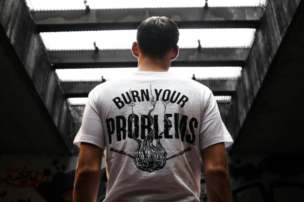 Man standing indoors, wearing a Graphic T-Shirt with 'Burn Your Problems' text, viewed from behind.
