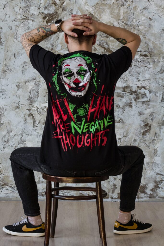 Man sitting on a chair, wearing a Joker-themed printed graphic t-shirt, hands on head